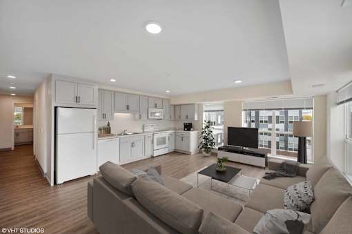 A modern living room with a grey sofa and a kitchen area in the background.