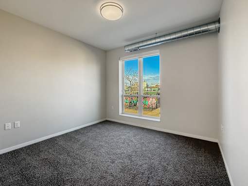A room with a grey carpet and a window showing a view of a tree and a building.