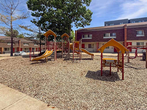 A playground with a yellow slide and a red and yellow play structure in front of a Liberty Plaza Townhome building.