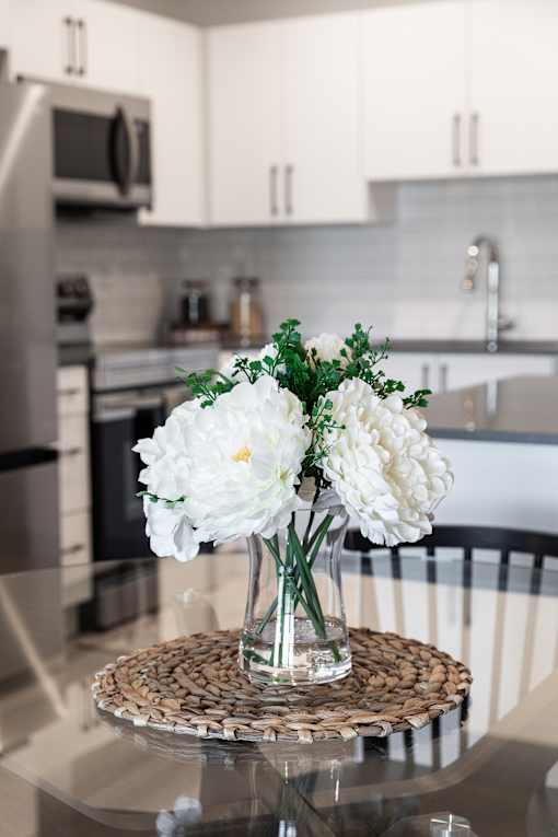 A glass vase with white flowers sits on a woven placemat on a glass table.