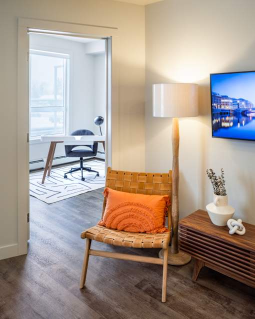 A chair with an orange pillow is in the foreground of a room with a television on the wall.