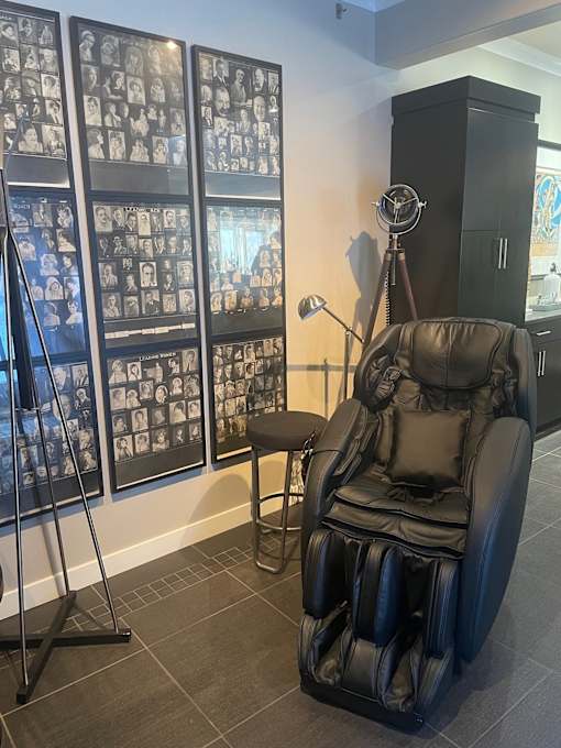 A black leather massage chair sits in front of a wall of framed photos.