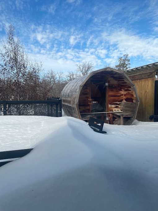 A wooden barrel sauna sits in the snow in front of a wooden shed.