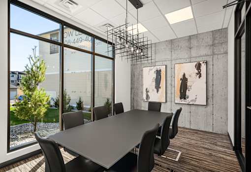 A conference room with a long table and chairs, a window with a view of a building and a tree, and two paintings on the wall.