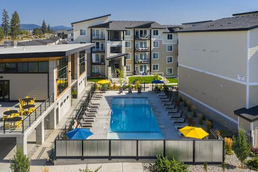 A large swimming pool surrounded by lounge chairs and umbrellas in front of apartment buildings.