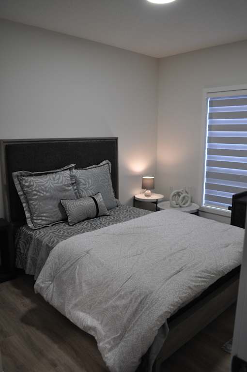 A bed with a grey comforter and pillows is in a room with a window and a lamp at The Laurent at Laurel Apartments, Edmonton, AB, T6T 2T5