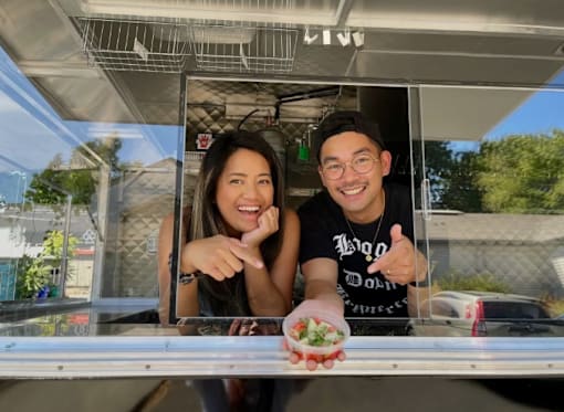 A man and woman are standing behind a food truck counter, smiling and pointing at the camera.