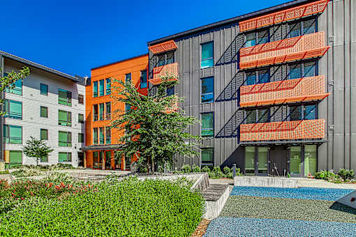 A modern building with orange balconies is surrounded by greenery and a small pond.