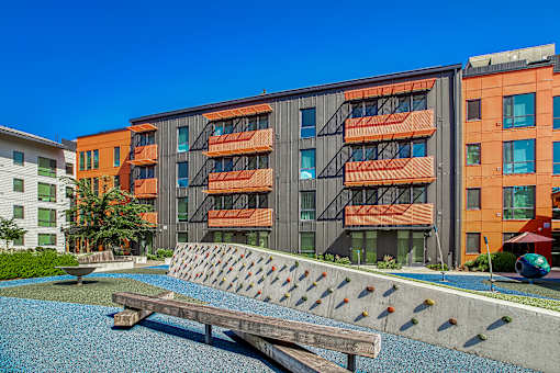 A modern building with orange and grey balconies is surrounded by a small pool and a wooden bench.
