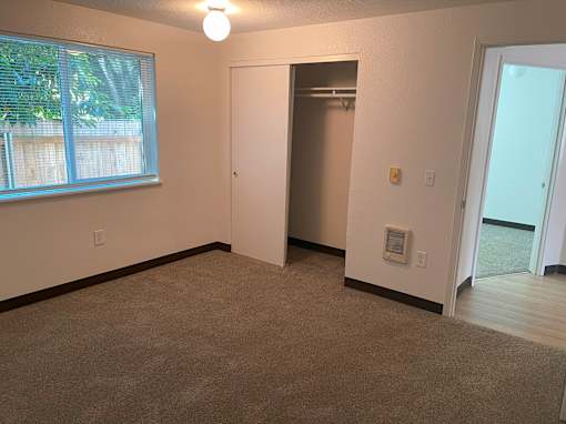 A room with a carpeted floor, a window with blinds, a white door, and a light switch on the wall.