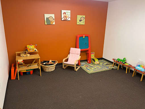 A room with orange walls and a grey carpet has a wooden chair, a table, and a basket on the floor.