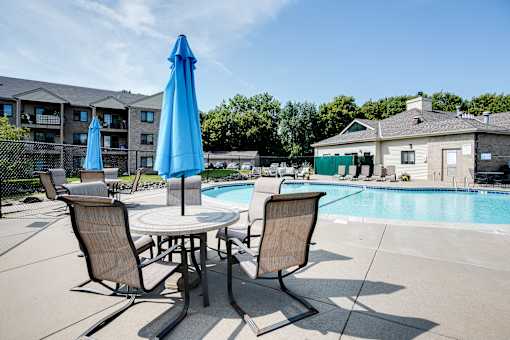 A table with two chairs and a blue umbrella sits by a pool.