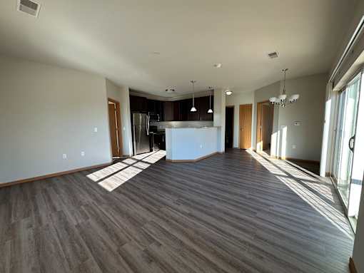 A spacious room with wooden flooring and a kitchen area in the background.
