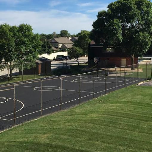 A basketball court is surrounded by a fence and there are houses in the background.