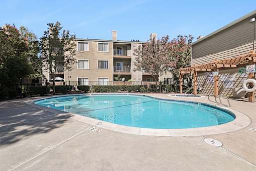 Resort-style swimming pool at Caifornia Place in gated pool area surrounded by lush greenery and blue skies for a beautiful and relaxing poolside experience. With plenty of seating including dining tables and chaise lounges