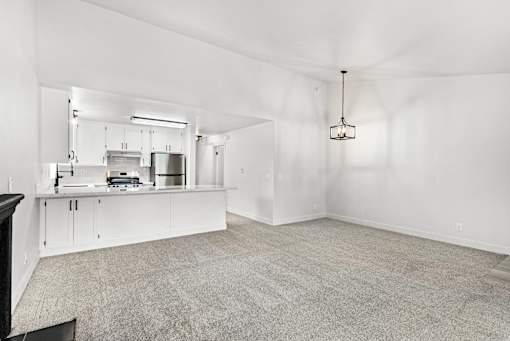 A spacious kitchen with white cabinetry and living area with and neutral carpet.
