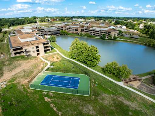 A tennis court is located on a grassy area next to a lake. at The Residences at Tesson Ridge Apartments, Saint Louis, MO, 63128