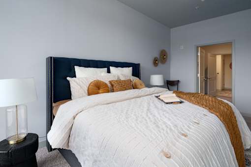 A bed with a white comforter and a blue headboard with a gold pillow and a book on it. at The Residences at Tesson Ridge Apartments, Missouri