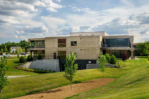 A modern building with a flat roof and large windows is surrounded by a grassy area with trees at The Residences at Tesson Ridge Apartments, Saint Louis, MO