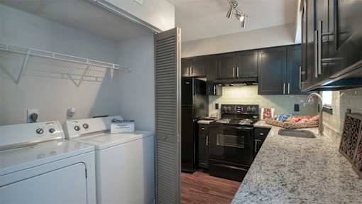 Duluth, GA Townhomes - Amberlake Village - Laundry Closet With Full Sized Washer And Dryer Beside Modern Kitchen With Dark Cabinets And Granite Countertops.
