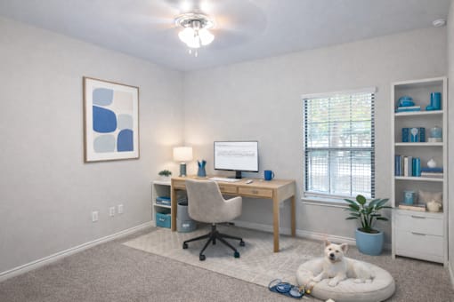A room with a desk, chair, and a dog on a bed.