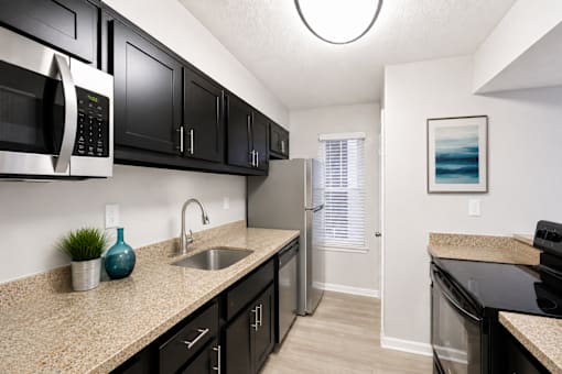 One-Bedroom Apartments in Sandy Springs, GA - Veridian at Sandy Springs - Kitchen With Black Cabinets, Beige Countertops, Stainless Steel Appliances, A Single Basin Sink, And Light Wood Flooring.