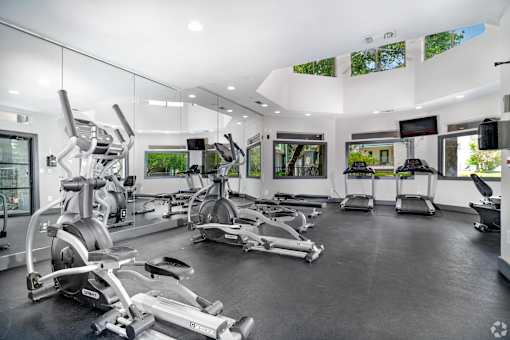 Churchill Estates, TX, Apartments Near San Antonio International Airport - Apex Apartments - Fitness Center With Elliptical Machines, Treadmills, Mirror Wall, Large Windows, and Black Flooring.