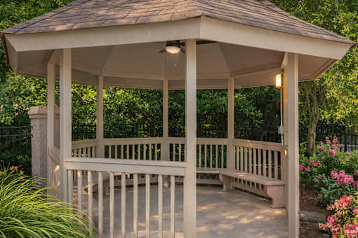 A gazebo with a brown roof and white pillars is surrounded by greenery and flowers.