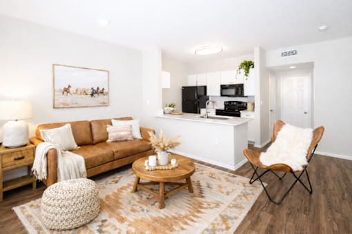 Pet-Friendly Apartments in Churchill Estates, TX - Apex Apartments - Living Room With A Brown Sofa, Hardwood Floor, Patterned Rug, Accent Chair, And Open Kitchen With Black Appliances.