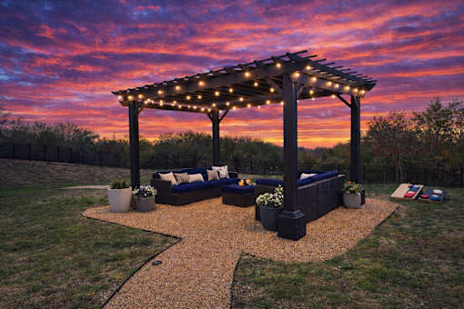 A gazebo with string lights is surrounded by a gravel path and potted plants.
