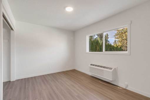 A room with a window showing trees and a white air conditioner on the floor.
