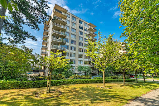 A tall apartment building with balconies is surrounded by green trees and bushes.