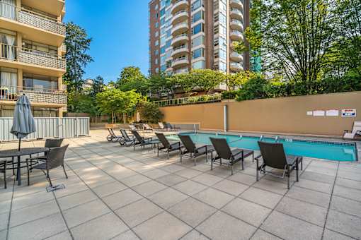 A pool area with chairs and a table is surrounded by buildings and trees.