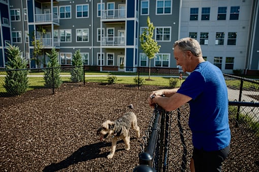 A man in a blue shirt is standing on a gravel area with a small dog.
