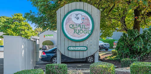 A sign for a Quail Ridge is in front of a white fence and green bushes.