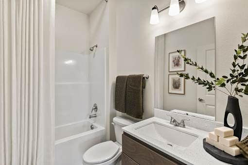 A bathroom with a white toilet, a white sink, a white bathtub, a mirror, a plant, and a towel.