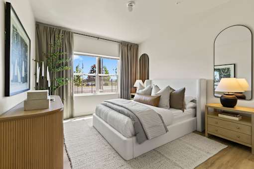 A bedroom with a large bed, a wooden nightstand, and a large window with curtains.