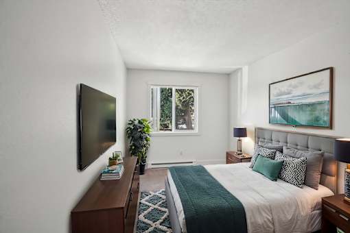 A bedroom with a bed, a television, a window, and a painting on the wall.
