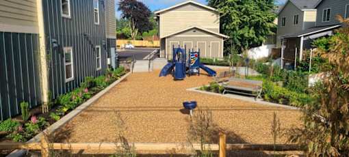A playground with a slide and a bench is surrounded by a fence and some houses.