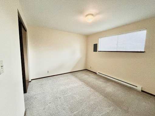 A room with a carpeted floor, a window with blinds, and a small speaker mounted on the wall.