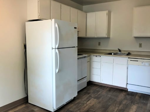 A white refrigerator stands in a kitchen with white cabinets and a wooden floor.