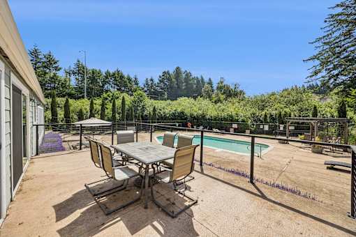 A table and chairs are set up on a patio with a pool and trees in the background.