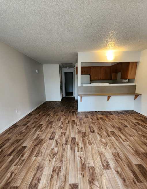 A room with wooden flooring and a kitchen area in the background.