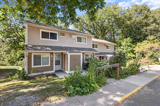 A two-story house with a brown and beige exterior is surrounded by greenery.