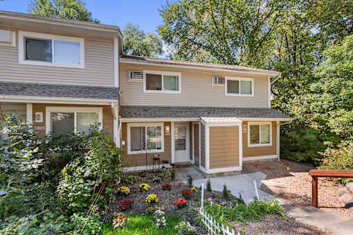 A house with a brown and beige exterior is surrounded by greenery and has a small garden in front.