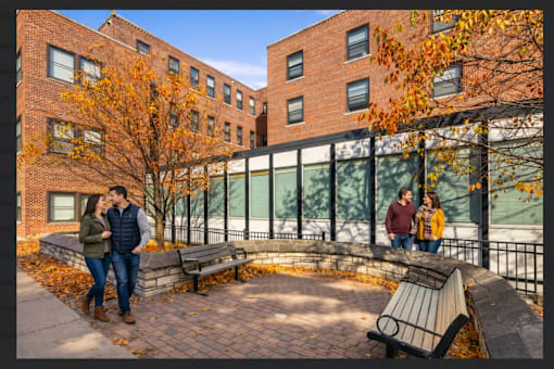 A couple is standing on a brick patio in front of a brick building with a tree with orange leaves.