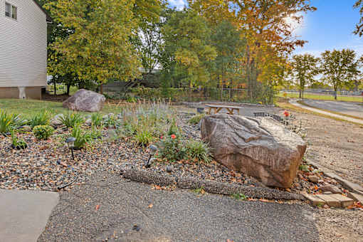 A large rock sits in the middle of a gravel area with a grassy area and trees in the background.
