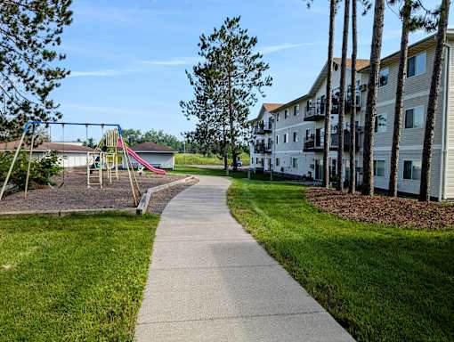 A playground with a slide and swings is surrounded by a grassy area and a sidewalk.
