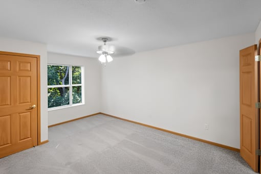 A room with a carpeted floor, a ceiling fan, and a window with a view of trees outside.
