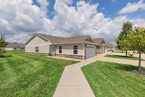 A house with a brown roof and a grey garage door is surrounded by a grassy lawn and a sidewalk.